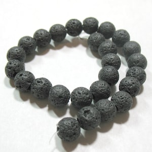 May include: Strand of black lava rock beads. The beads are round, with a porous, textured surface. The beads are strung on a white string, ready for jewelry making. Each bead is approximately 1 cm in diameter.