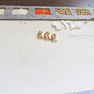 May include: A white felt beading mat with a gray border and compartments for beads. The mat has a white and gold beaded design in the center, with white beads scattered around it. There is a needle and thread on the mat.