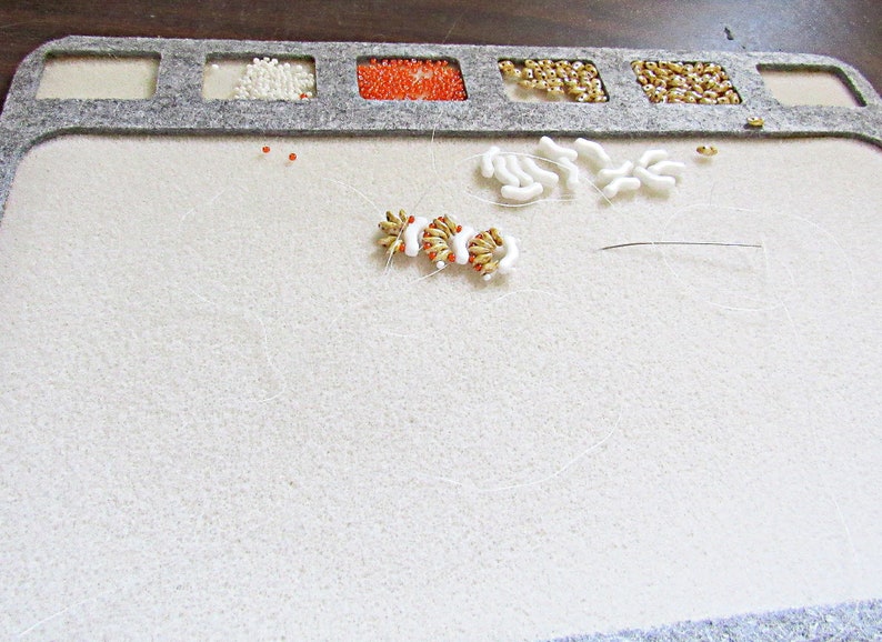 May include: A white felt beading mat with a gray border and compartments for beads. The mat has a white and gold beaded design in the center, with white beads scattered around it. There is a needle and thread on the mat.