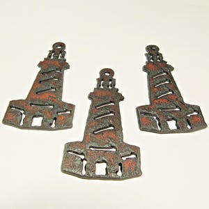 May include: Three decorative lighthouse cutouts with a rustic, weathered appearance. The metal cutouts are primarily dark brown with reddish-brown accents, featuring horizontal slits and square openings. The lighthouses have a textured surface and are arranged on a white background.