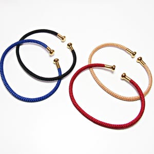 May include: Four open-ended cuff bracelets featuring a textured, braided cord design in black, blue, red, and beige. Each bracelet is finished with polished gold-tone metal caps at both ends, creating a simple and uniform circular shape.