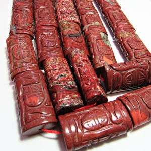May include: Close-up of a strand of carved red jasper beads. The beads are cylindrical with intricate relief carvings. The color is a deep red with some variations in the stone. The beads are strung on a red cord, suggesting they are for jewelry making.
