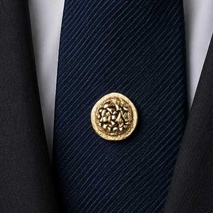 May include: A gold-colored tie pin with a detailed, circular design is centered on a navy blue striped tie. The tie is knotted and set against a white collared shirt and a dark suit jacket.
