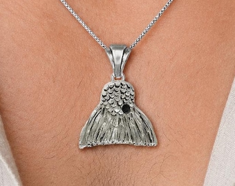 May include: A silver-toned pendant shaped like a fish tail, featuring a textured scale pattern and a small, dark circular accent at the top. The pendant hangs from a thin, metallic box chain necklace that rests against a light-colored collared shirt.