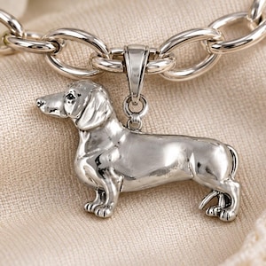 May include: A silver-toned metal pendant shaped like a dachshund dog, suspended from a thick oval-link chain. The pendant features a detailed, smooth finish with visible facial features and paws. The text "Chain sold separately." is visible.