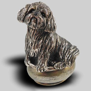 Op de afbeelding: Een zilveren beeldje van een Shih Tzu hond die in een kom zit. De hond is gedetailleerd met vacht en heeft een realistische uitdrukking.