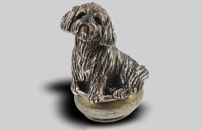 Op de afbeelding: Een zilveren beeldje van een Shih Tzu hond die in een kom zit. De hond is gedetailleerd met vacht en heeft een realistische uitdrukking.