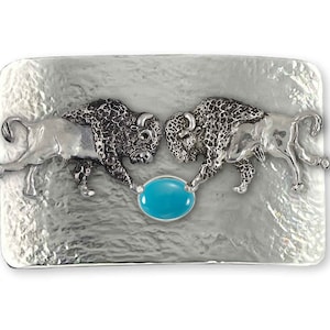 May include: Silver belt buckle featuring two buffalo facing each other, with a turquoise stone in the centre.