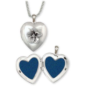 Pode incluir: Um colar com um pingente em forma de coração prateado com um design floral. O medalhão abre para revelar dois espaços com um fundo azul. O colar é exibido em um fundo branco.