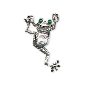 Peut inclure: Un charme de grenouille en argent avec des yeux verts, suspendu à une chaîne. La grenouille est en position d'escalade, les pattes tendues.
