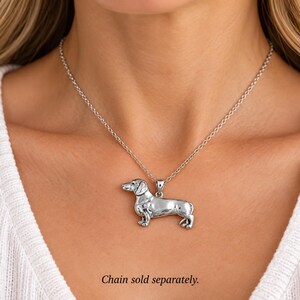 May include: A silver-toned metal pendant shaped like a dachshund dog, featuring a smooth, polished finish and detailed anatomical features. The pendant is suspended from a delicate silver-colored chain. The text Chain sold separately is visible.