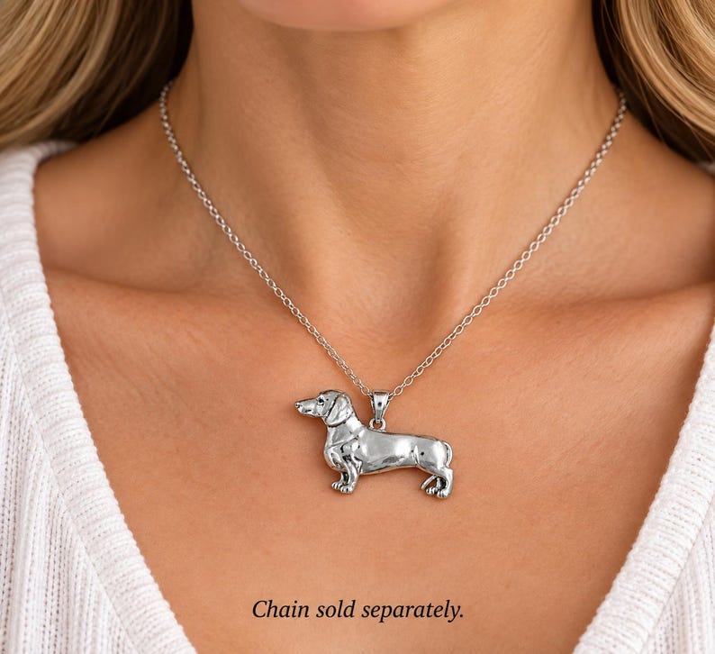 May include: A silver-toned metal pendant shaped like a dachshund dog, featuring a smooth, polished finish and detailed anatomical features. The pendant is suspended from a delicate silver-colored chain. The text Chain sold separately is visible.