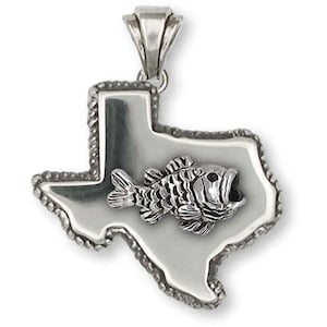 Puede incluir: Colgante de plata con la forma del estado de Texas, con un diseño detallado de un pez en el centro. El colgante tiene un borde en forma de cuerda y una argolla para sujetar a una cadena. El pez mira hacia la izquierda con la boca abierta.