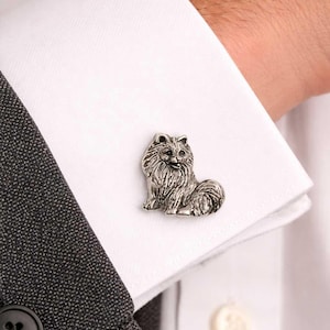 Pode incluir: Uma abotoadura prateada em forma de cão da raça Pomerânia, pousada na manga de uma camisa branca. A abotoadura é detalhada com pelos e traços faciais. A camisa é combinada com um casaco cinzento.