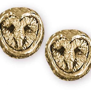 May include: Two gold-colored owl-shaped stud earrings. Each earring features a detailed owl face design with textured features. The earrings are set against a white background, highlighting their metallic sheen and intricate craftsmanship.