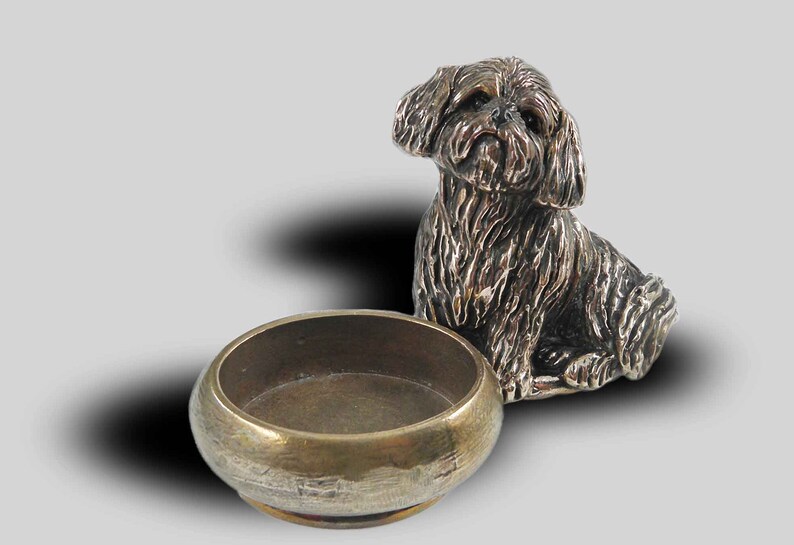 Op de afbeelding: Een zilveren beeldje van een kleine, harige hond die naast een kleine, ronde messing kom zit.