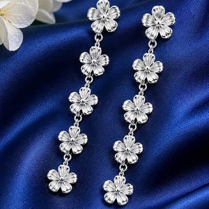 May include: Silver-toned dangle earrings featuring a floral design. Each earring has five flower-shaped charms connected by delicate chains. The earrings are displayed against a dark blue satin background, with a white flower in the upper left corner.