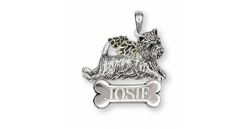 May include: Silver pendant with a detailed, three-dimensional image of a Cairn Terrier with gold wings. The dog is lying on a bone-shaped charm with the name "JOSIE" engraved on it.