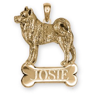 May include: A gold-colored pendant in the shape of a dog, standing above a bone-shaped tag. The tag is inscribed with the name "JOSIE". The dog pendant has a detailed, textured appearance, and a loop for a chain.