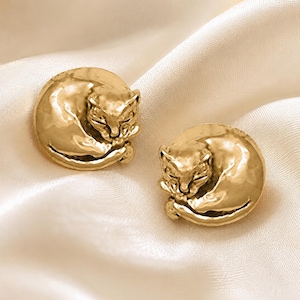 May include: A pair of gold-toned stud earrings, each featuring a circular design with a sculpted, curled-up cat in a resting position. The metal surface has a polished, reflective finish that catches the light, highlighting the detailed feline features.