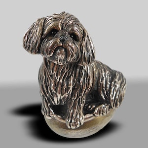 Op de afbeelding: Een zilverkleurig beeldje van een Shih Tzu hond die op een ronde basis zit. De hond is afgebeeld met een pluche vacht en een lieve uitdrukking.