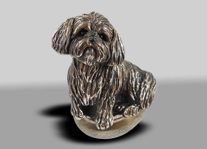 Op de afbeelding: Een zilverkleurig beeldje van een Shih Tzu hond die op een ronde basis zit. De hond is afgebeeld met een pluche vacht en een lieve uitdrukking.