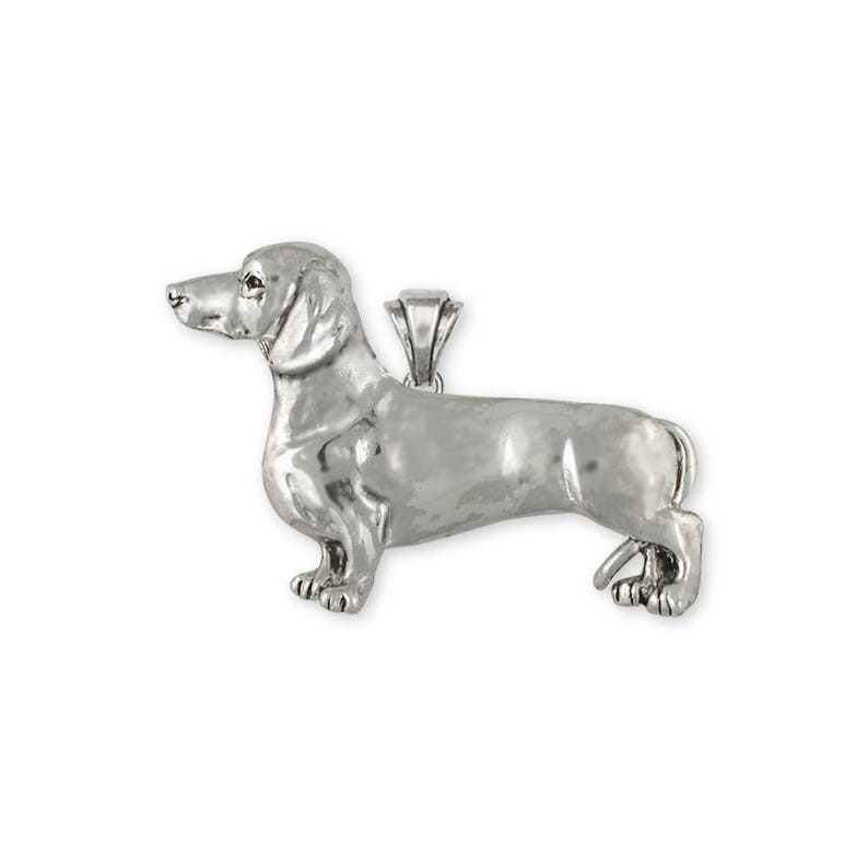 May include: A silver-toned metal pendant shaped like a dachshund dog in a standing profile position. The pendant features a smooth, reflective surface with detailed contours of the dog's head, long body, and short legs, including a small bail at the top.