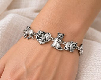 May include: A silver-toned metal link bracelet featuring a series of detailed cat charms. The charms depict cats in various poses, including one cat resting on a heart-shaped base, all connected by small circular metal jump rings.