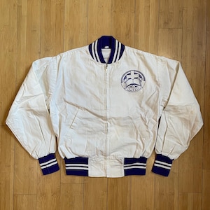 May include: A white varsity jacket with blue and white striped ribbing around the collar, cuffs, and waistband. The jacket has a zipper closure and a large embroidered patch on the front that says "LIBIA" with a cross and an open book.