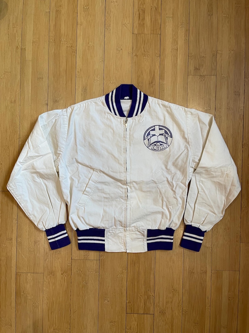 May include: A white varsity jacket with blue and white striped ribbing around the collar, cuffs, and waistband. The jacket has a zipper closure and a large embroidered patch on the front that says "LIBIA" with a cross and an open book.