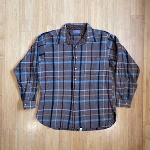 以下が含まれることがあります： ボタン留めのフロントと長袖の、茶色、青、タンの格子柄のフランネルシャツ。シャツは左胸にパッチポケットと、コントラストの効いた青い裏地の襟が付いている。