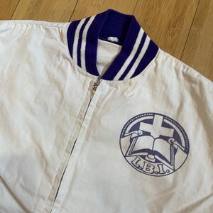May include: White cotton bomber jacket with a purple and white striped collar and a purple logo on the front. The logo is a circle with a cross, a book, and the letters "L.B.I."