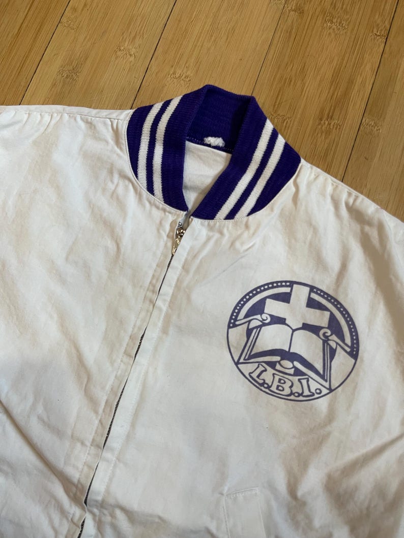 May include: White cotton bomber jacket with a purple and white striped collar and a purple logo on the front. The logo is a circle with a cross, a book, and the letters "L.B.I."