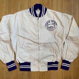 May include: A white varsity jacket with a blue and white striped collar and cuffs. The jacket has a zipper closure and a patch on the left chest with a cross and a book. The patch also has the text "L.B.J.A. 1963" on it.