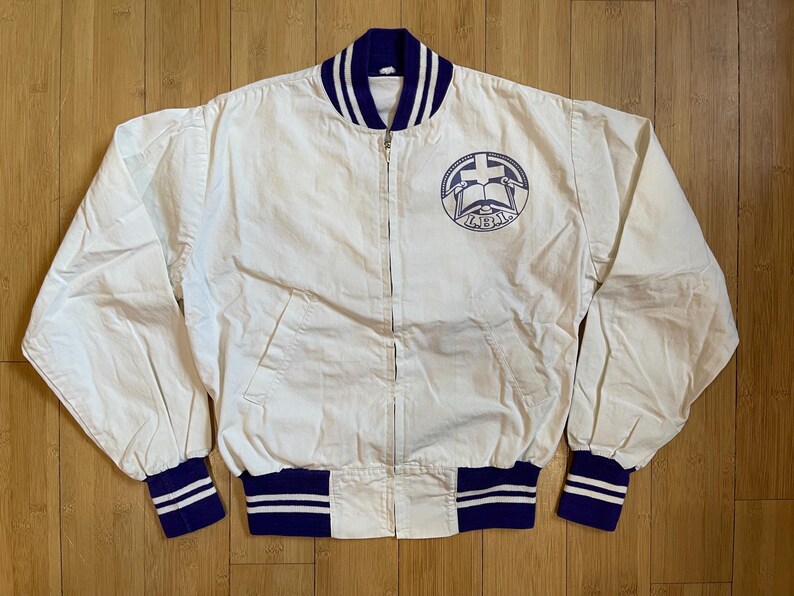May include: A white varsity jacket with a blue and white striped collar and cuffs. The jacket has a zipper closure and a patch on the left chest with a cross and a book. The patch also has the text "L.B.J.A. 1963" on it.