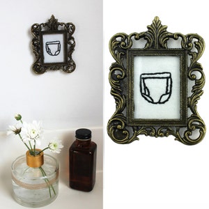 Op de afbeelding: Een antiek ogend bronzen frame met een geborduurd luierontwerp op witte stof. Het frame is versierd en hangt aan een witte muur. Een kleine vaas met witte bloemen en een bruine fles staan op de voorgrond. Het frame heeft een sierlijk ontwerp.