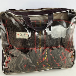 Puede incluir: Una bolsa de almacenamiento de plástico transparente con un asa de cuero marrón y una cremallera. La bolsa contiene una manta a cuadros con un patrón rojo, amarillo y negro. La manta tiene una etiqueta que dice "Farifax" y "Family Woven Blankets".