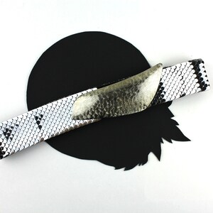Puede incluir: Un cinturón de piel de serpiente blanco y negro con una hebilla plateada. La hebilla es grande, curva y tiene una superficie texturizada.
