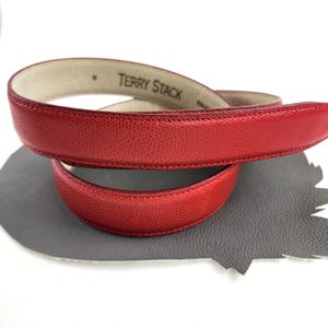 May include: A red leather belt with the text "TERRY STACK" embossed on the inside. The belt has a textured surface and is curled in a loop. The background is white and gray.