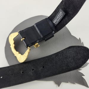 Puede incluir: Cintur&oacute;n de cuero negro con hebilla dorada. La hebilla tiene un dise&ntilde;o adornado y curvado. El cintur&oacute;n tiene una etiqueta que dice "BRIGHTON". El cintur&oacute;n est&aacute; sobre una superficie gris.