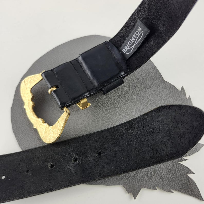 Puede incluir: Cintur&oacute;n de cuero negro con hebilla dorada. La hebilla tiene un dise&ntilde;o adornado y curvado. El cintur&oacute;n tiene una etiqueta que dice "BRIGHTON". El cintur&oacute;n est&aacute; sobre una superficie gris.