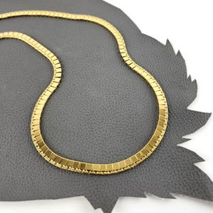May include: A gold-colored necklace with a flat, rectangular link design. The necklace is displayed on a gray surface. The chain is made of small, rectangular links that create a smooth, flexible band. The necklace is a classic jewelry piece.