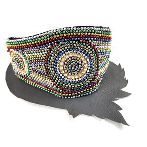 May include: A colourful beaded cuff bracelet with a circular design. The bracelet features red, green, blue, and gold beads arranged in concentric circles. The detailed pattern is set on a grey, leaf-shaped surface. The bracelet is a striking piece of jewellery.
