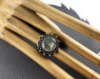 May include: A silver ring with a large, round, pale green stone. The ring has a detailed, beaded border around the stone. The ring is displayed on a wooden surface, with a dark gray background.