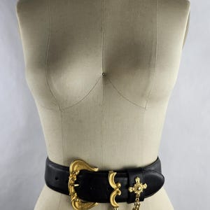 Puede incluir: Un cintur&oacute;n de cuero negro con una hebilla grande dorada y detalles decorativos. El cintur&oacute;n presenta una cadena dorada y un dije en forma de cruz. El cintur&oacute;n se muestra en un maniqu&iacute;.