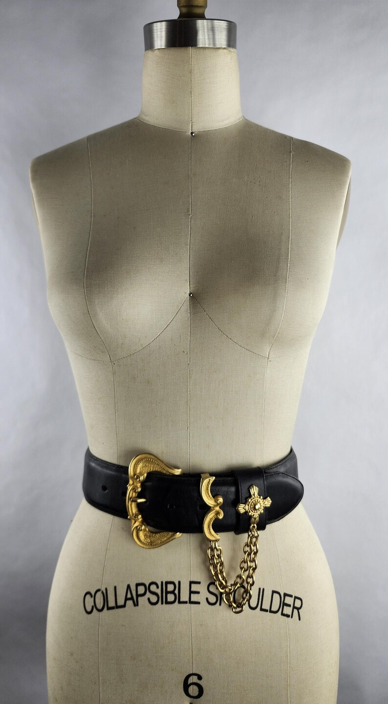 Puede incluir: Un cintur&oacute;n de cuero negro con una hebilla grande dorada y detalles decorativos. El cintur&oacute;n presenta una cadena dorada y un dije en forma de cruz. El cintur&oacute;n se muestra en un maniqu&iacute;.