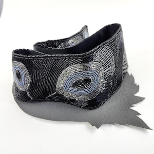 May include: A black beaded belt with a peacock feather design. The belt features intricate beadwork in shades of black, gray, and blue. The belt is placed on a gray leather base, creating a striking contrast.