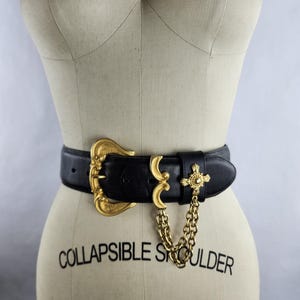 Puede incluir: Cintur&oacute;n de cuero negro con una banda ancha y una hebilla dorada ornamentada. El cintur&oacute;n presenta una cadena dorada decorativa y un adorno en forma de cruz. El maniqu&iacute; tiene la inscripci&oacute;n "COLLAPSIBLE SHOULDER" y el n&uacute;mero "6".