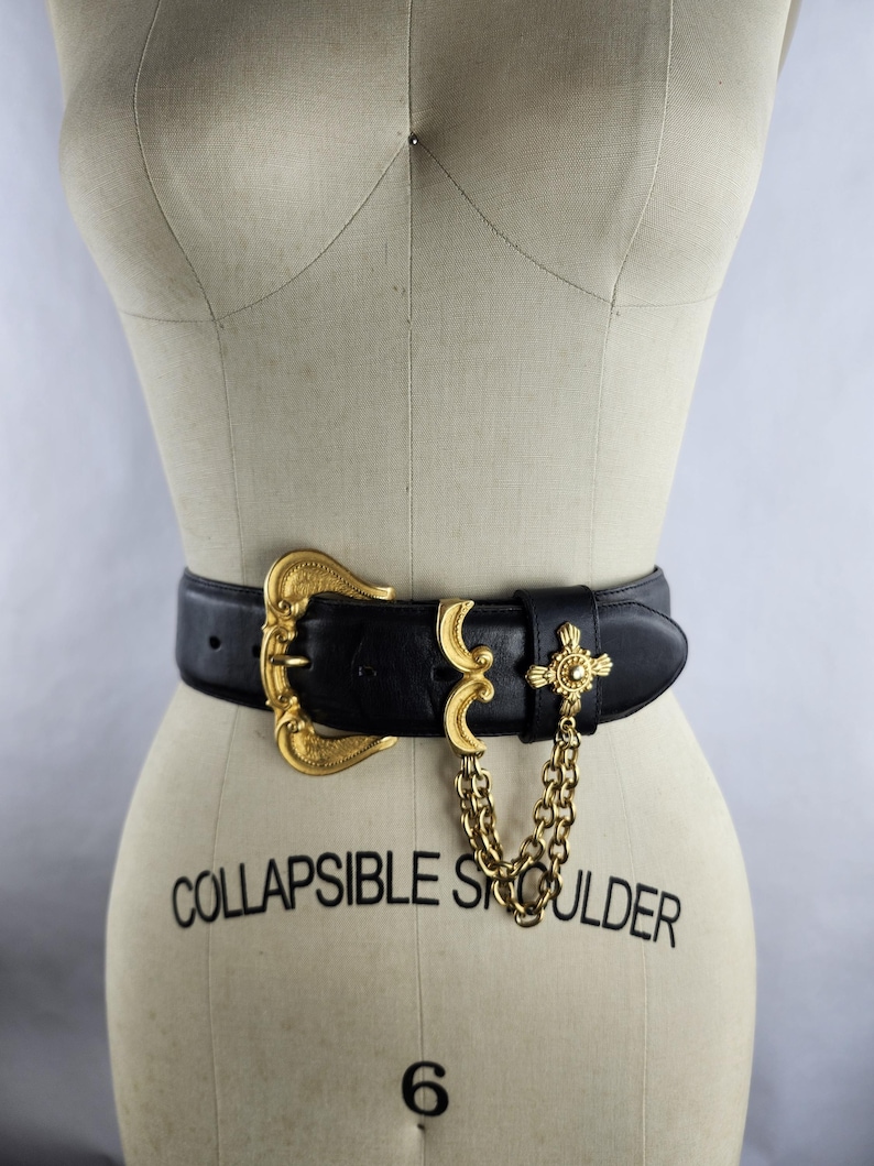 Puede incluir: Cintur&oacute;n de cuero negro con una banda ancha y una hebilla dorada ornamentada. El cintur&oacute;n presenta una cadena dorada decorativa y un adorno en forma de cruz. El maniqu&iacute; tiene la inscripci&oacute;n "COLLAPSIBLE SHOULDER" y el n&uacute;mero "6".