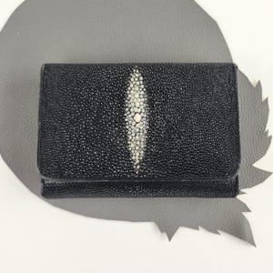 May include: A rectangular black wallet crafted from textured stingray leather. The exterior features a prominent, elongated, white, oval-shaped cluster of larger, smooth, bead-like scales centred vertically on the front flap of the wallet.